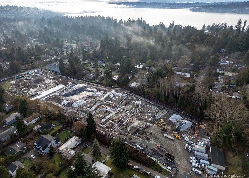 aerial view of a large construction site with equipment and trucks. there is a body of water in the background and houses in the foreground