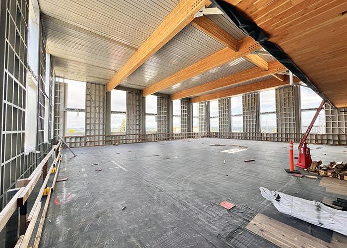 a large interior room under construction with part of the ceiling wood and large wood beams. Windows are in all three walls that are showing. The walls are framed but do not have wallboard.