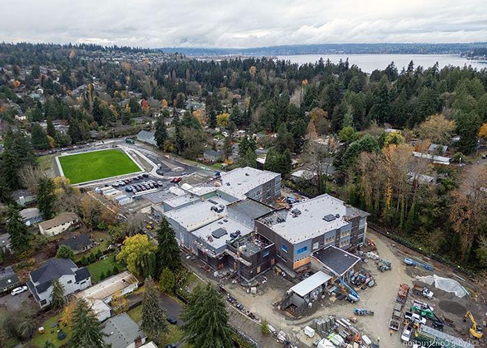 aerial of a large building under construction with a green field to the right and a lake in background